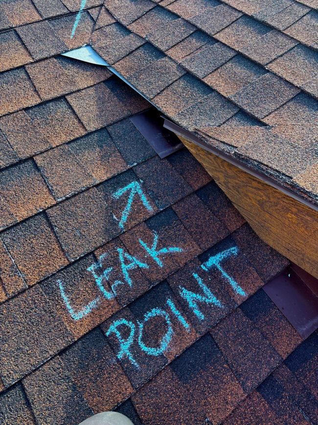 Leak point in a roof Asphalt shingle roof with “LEAK POINT” chalked pointing to faulty flashing at a roof-to-wall/valley intersection—illustrating a common Colorado roof leak that needs repair.