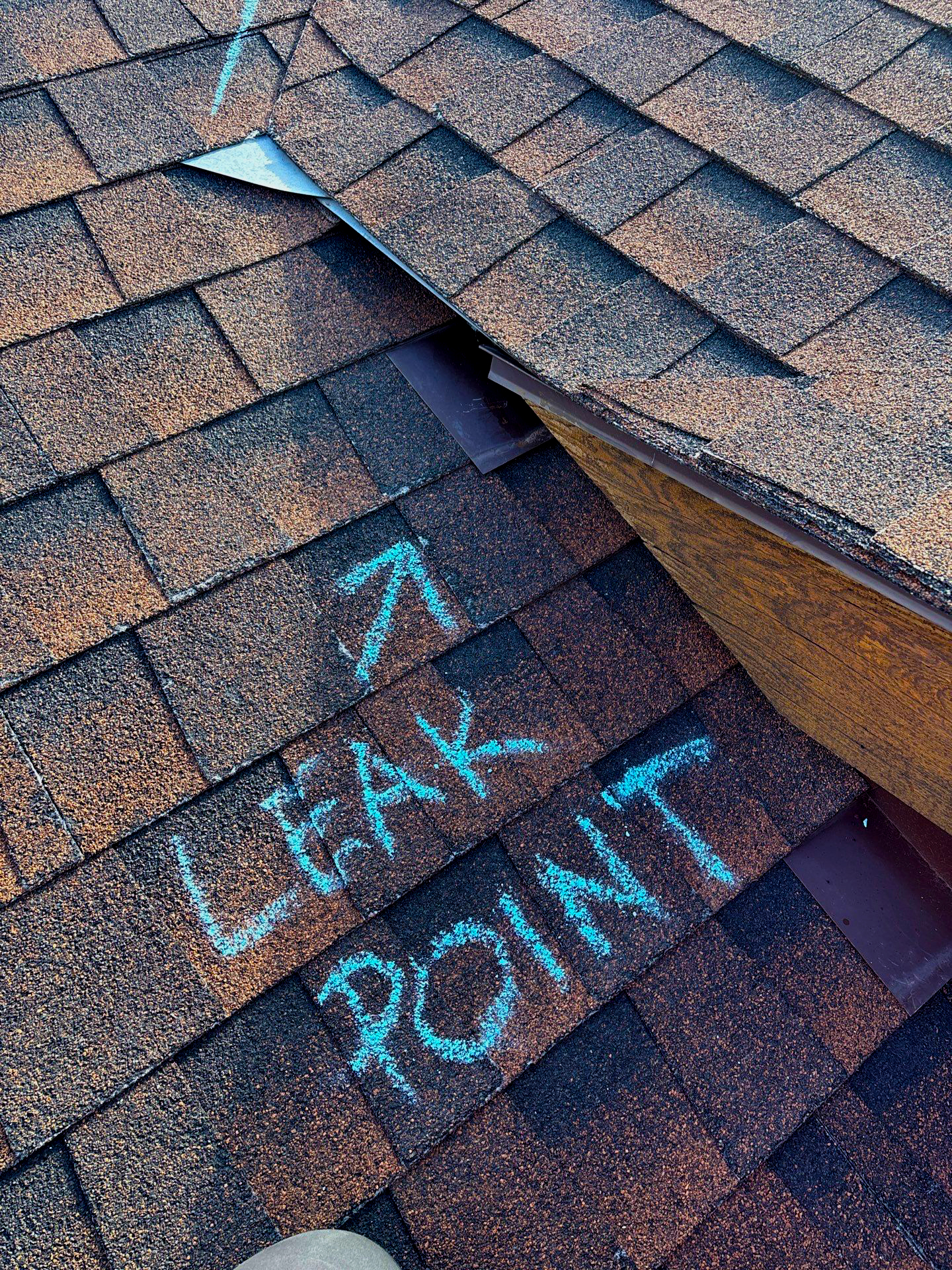 Asphalt shingle roof with “LEAK POINT” chalked pointing to faulty flashing at a roof-to-wall/valley intersection—illustrating a common Colorado roof leak that needs repair.