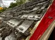 Old roof with severely damaged asphalt shingles, visible curling and cracking, and debris near the red wooden edge, showing signs of weather wear and urgent need for roof repair.