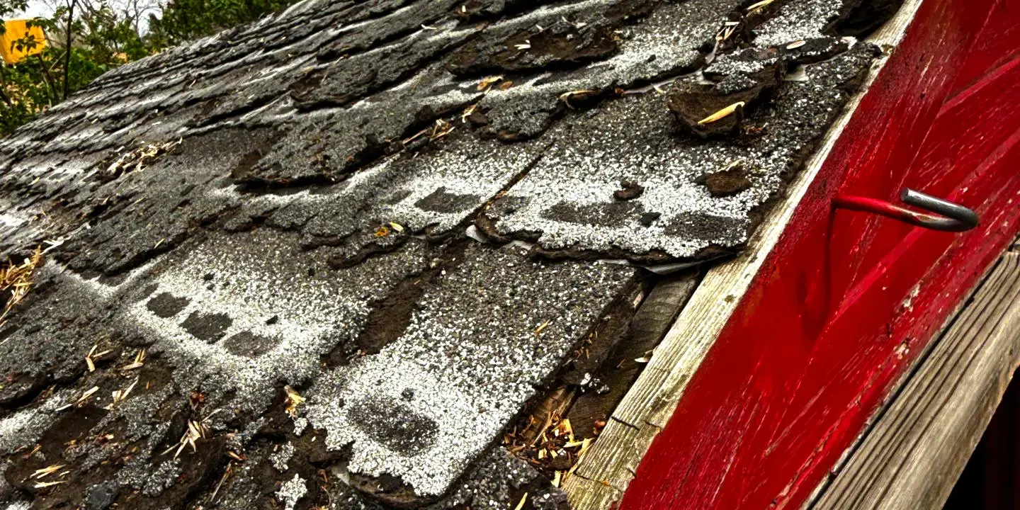 Old roof with severely damaged asphalt shingles, visible curling and cracking, and debris near the red wooden edge, showing signs of weather wear and urgent need for roof repair.