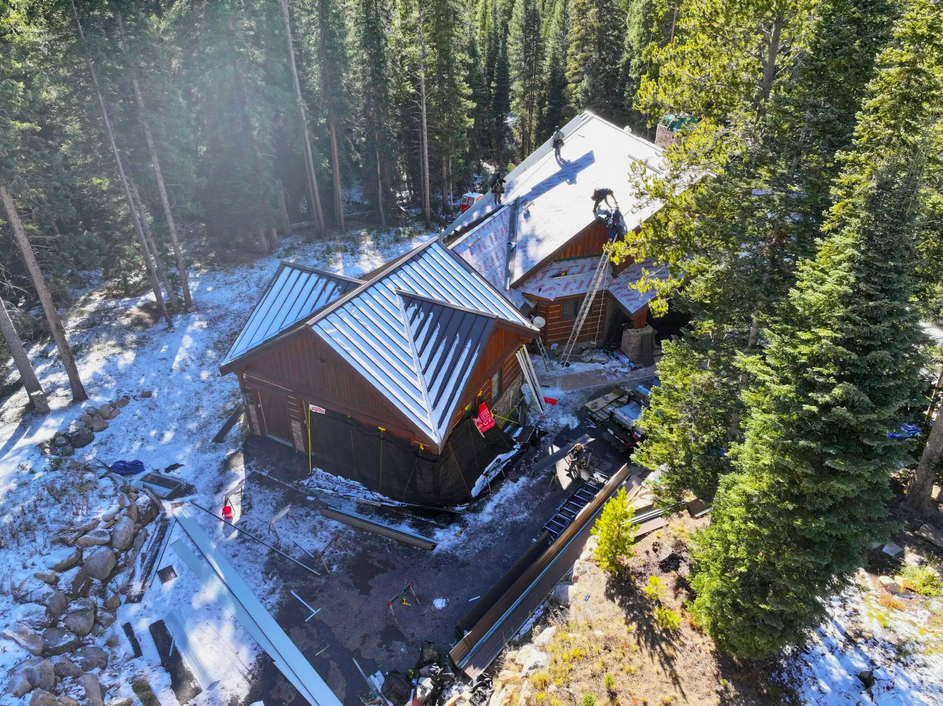 Roofing crew working on the installation of a metal roof on a house surrounded by trees and light snow.