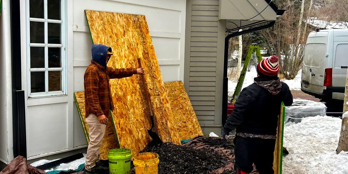 Roofing crew performing residential roof repair during winter, removing shingles and protecting the home with plywood and tarps in Colorado.