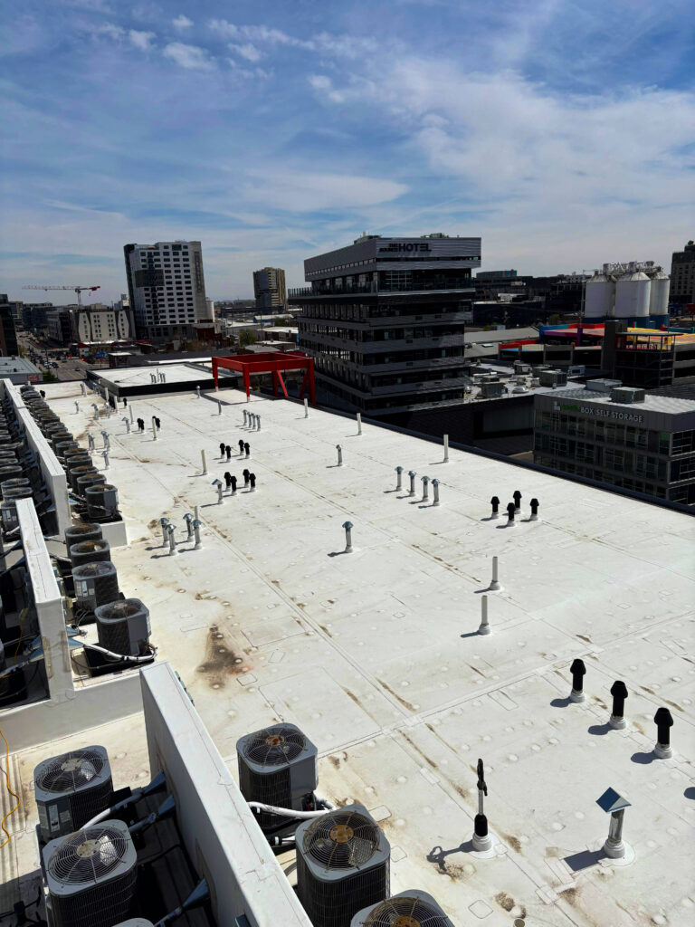 Flat commercial roof with white membrane, HVAC units, vents, and city buildings in the background under a blue sky.