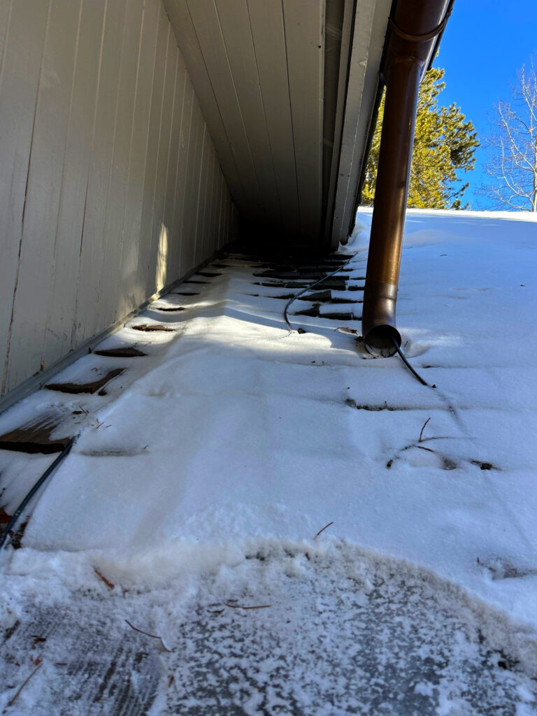Snow-covered roof valley with exposed shingles, heat cable, and downspout showing winter ice dam prevention on a residential roof.