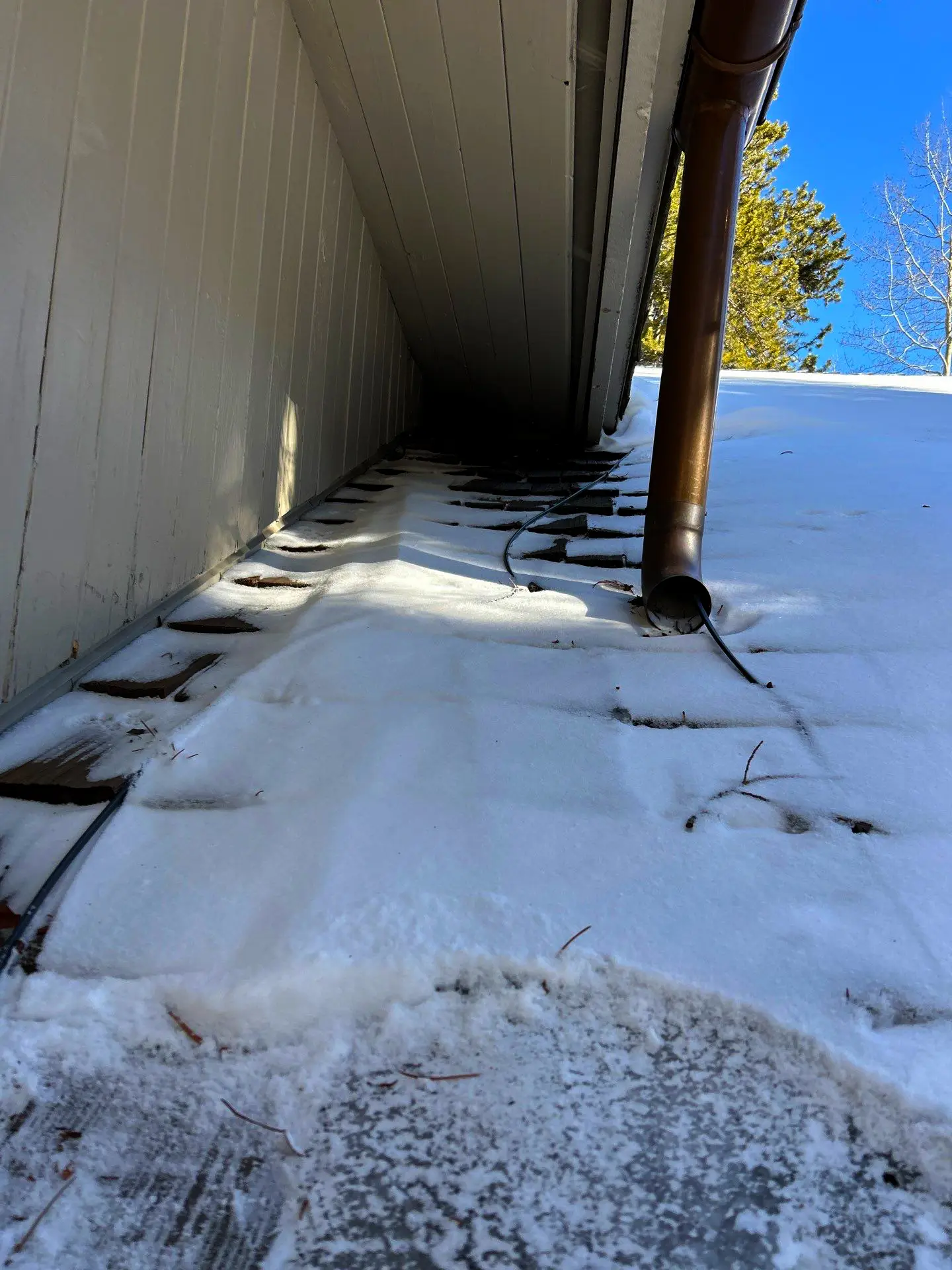 Snow-covered roof valley with exposed shingles, heat cable, and downspout showing winter ice dam prevention on a residential roof.
