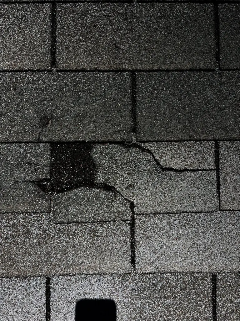 damaged asphalt shingles with cracks, granule loss, and a dark moisture stain indicating the need for emergency roof repair.