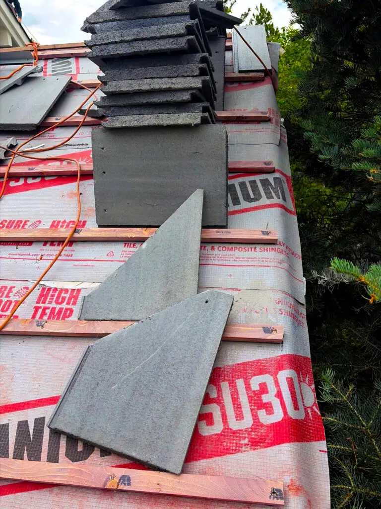 Roof installation in progress with synthetic underlayment, wood battens, and gray roofing tiles stacked on a sloped residential roof.