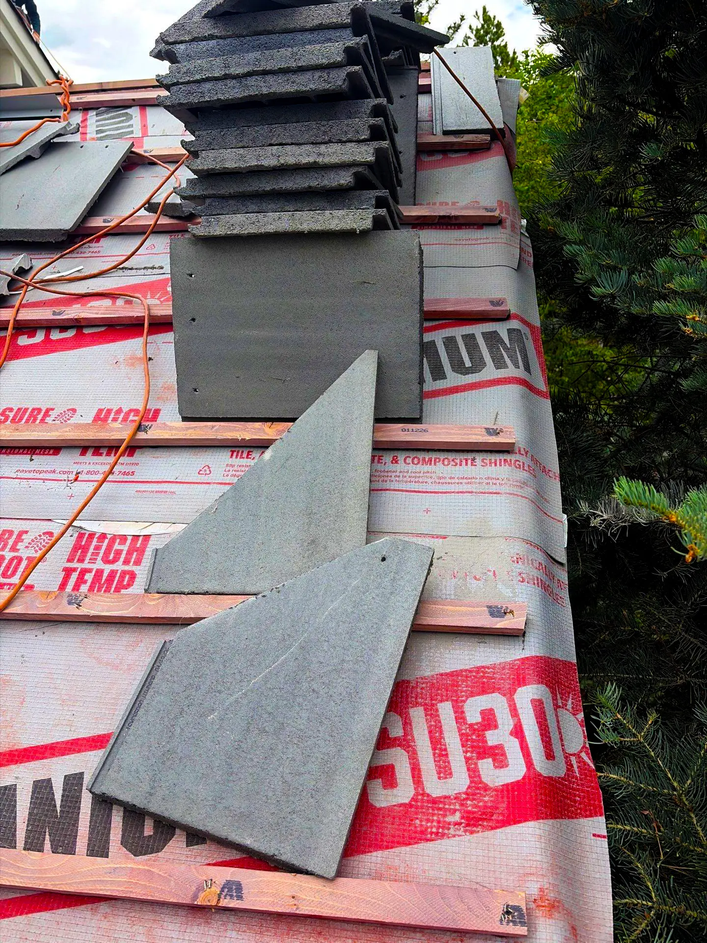Roof installation in progress with synthetic underlayment, wood battens, and gray roofing tiles stacked on a sloped residential roof.
