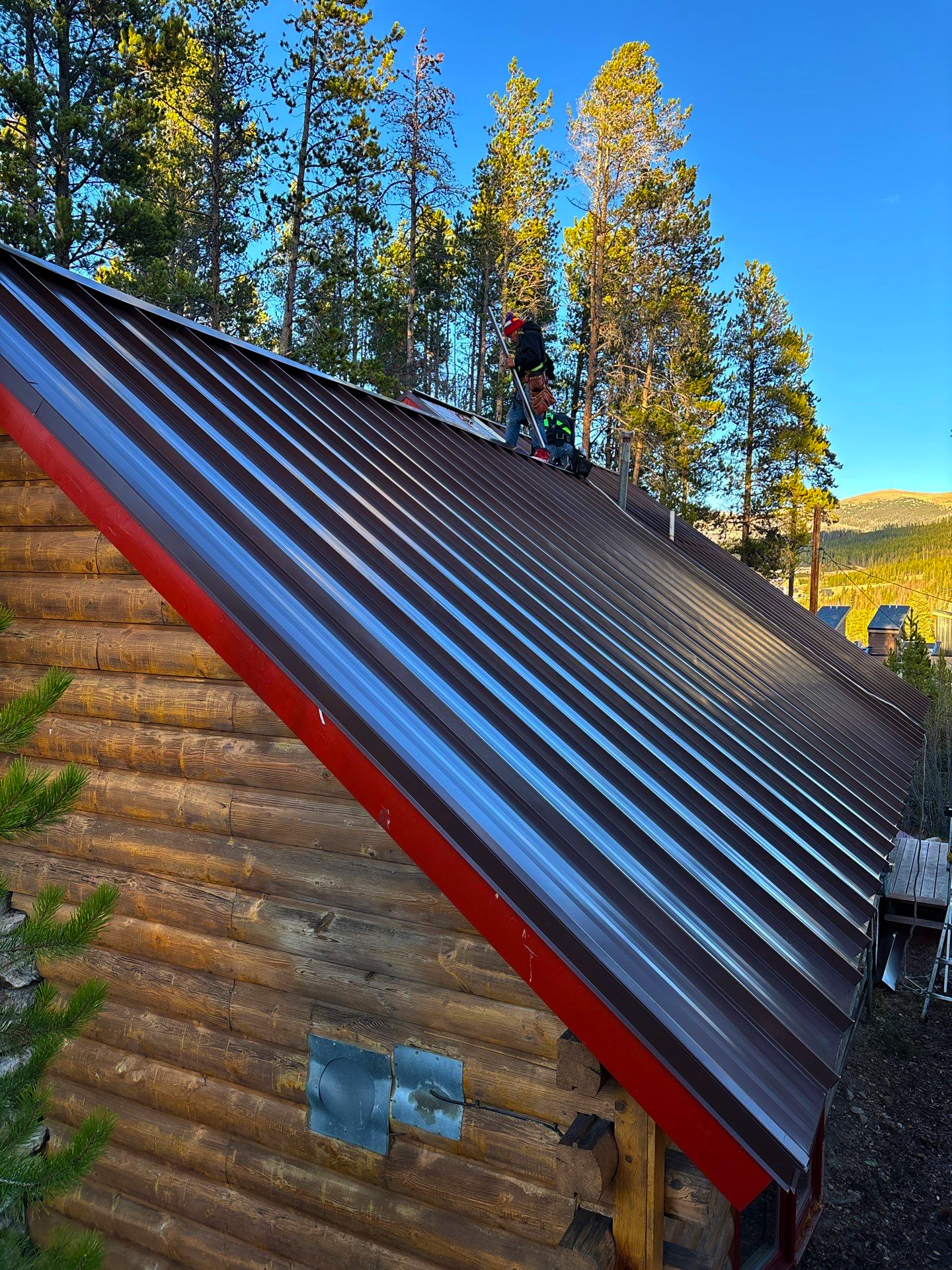 Roofer installing a dark metal roof on a wooden mountain cabin surrounded by pine trees in Colorado.